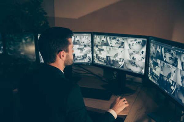 A man in a suit looks over some computer monitors that show various camera feeds